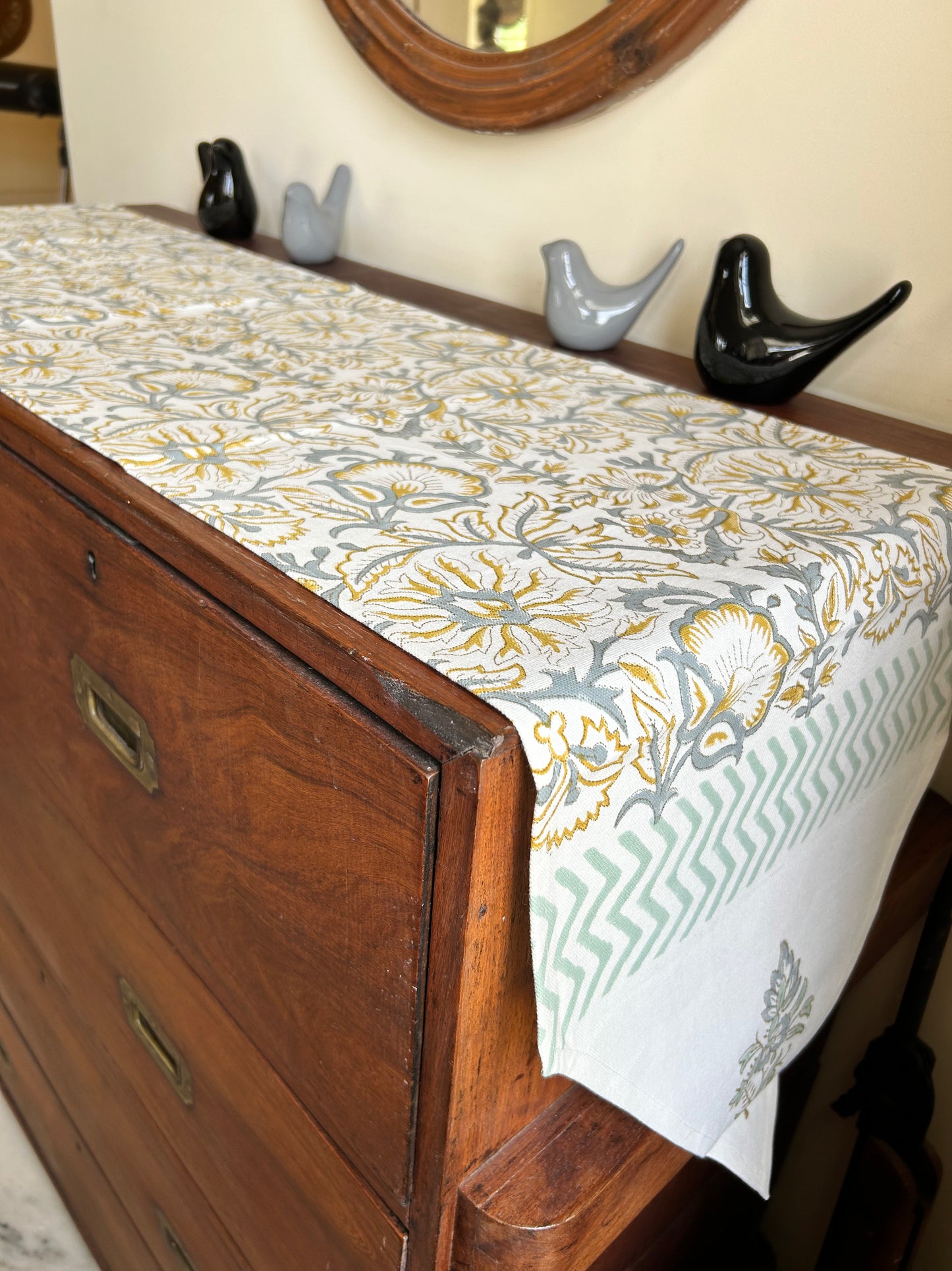 Wooden dresser with a patterned table runner draped over it, decorative birds on a shelf in the background.